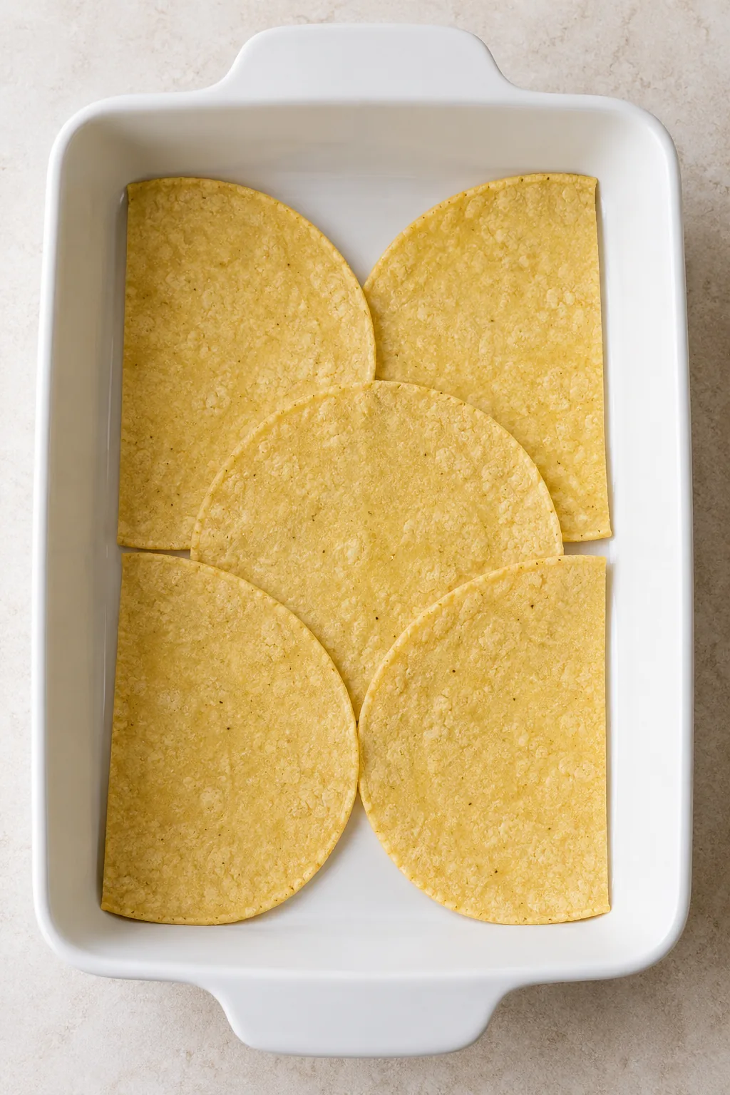 Corn tortillas arranged in the bottom of a white rectangular baking dish
