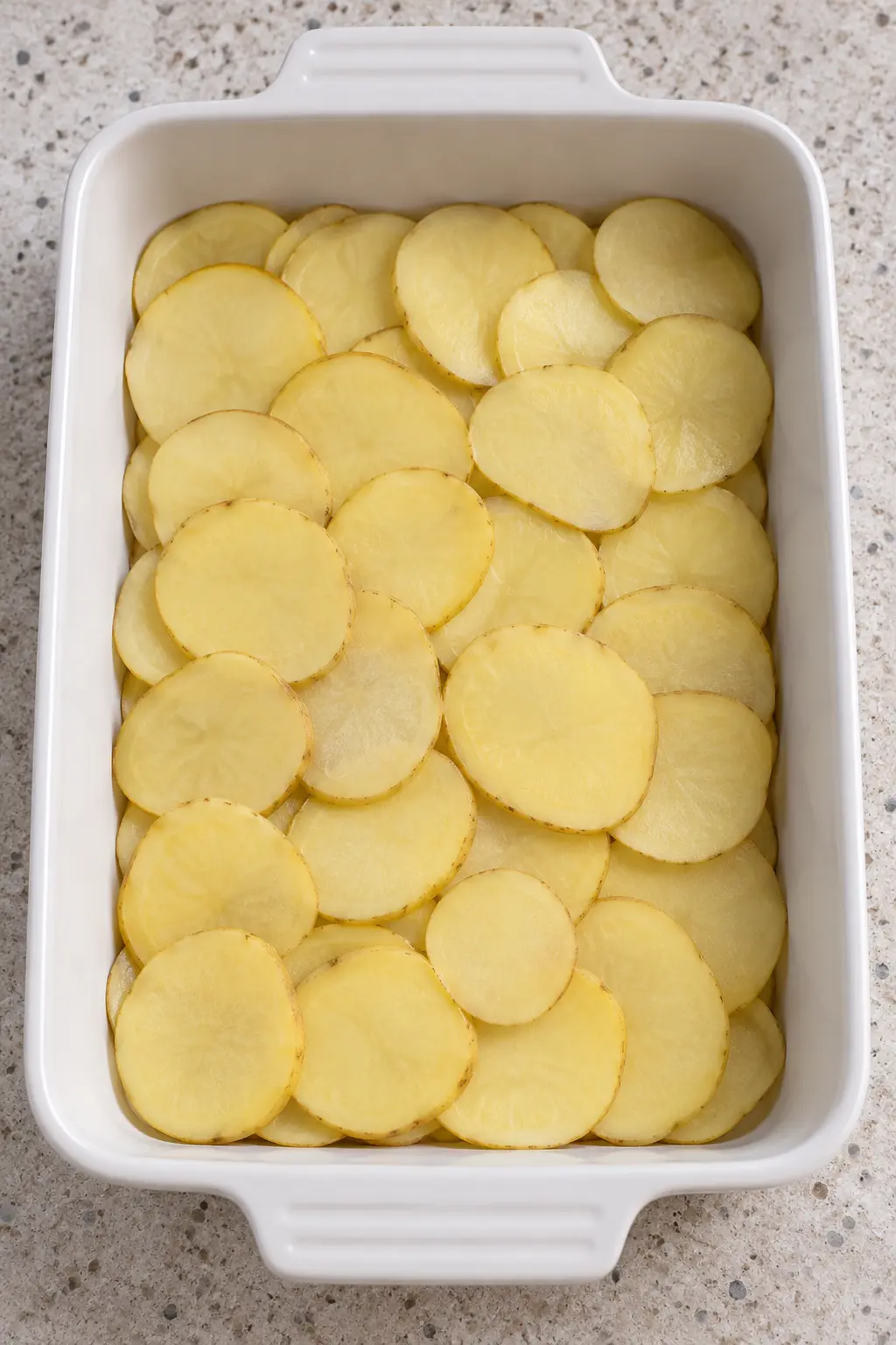 First layer of sliced potatoes in a white baking dish