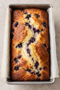 Golden blueberry cream cheese loaf cooling in its parchment-lined pan