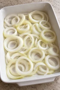 Raw sweet onion rings spread across the bottom of a white baking dish
