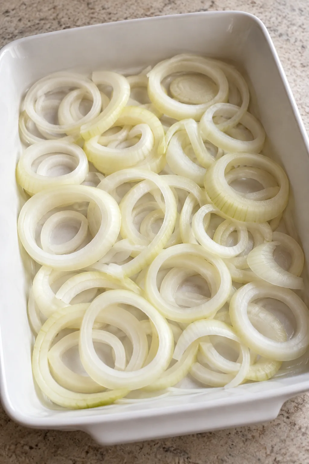 Raw sweet onion rings spread across the bottom of a white baking dish