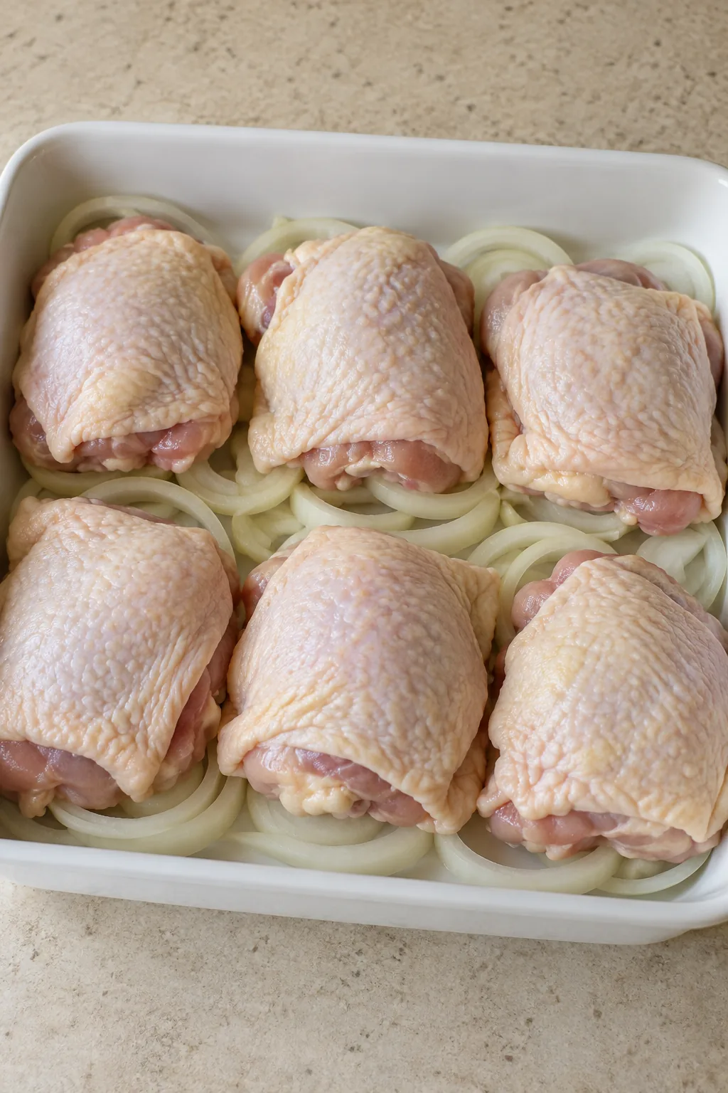 Six raw skin-on chicken thighs in one layer over sweet onion rings in a white baking dish