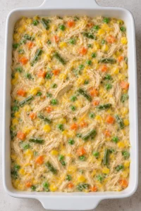 Creamy shredded chicken and mixed vegetable filling spread in a white casserole dish before adding biscuits
