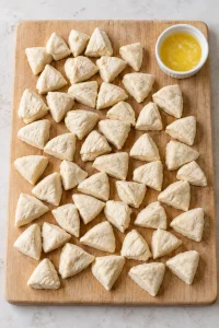 Raw biscuit dough cut into quarter pieces on a wooden board beside melted butter