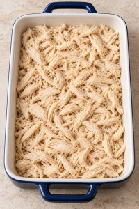 Cooked shredded chicken spread across the bottom of a white-interior navy baking dish