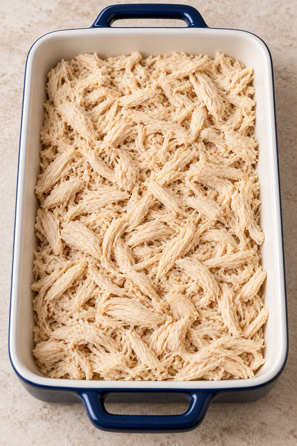 Cooked shredded chicken spread across the bottom of a white-interior navy baking dish