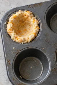 Cheddar biscuit dough pressed into a cup in a greased muffin well