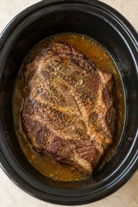 A seared chuck roast with seasoned cooking liquid in a black slow cooker