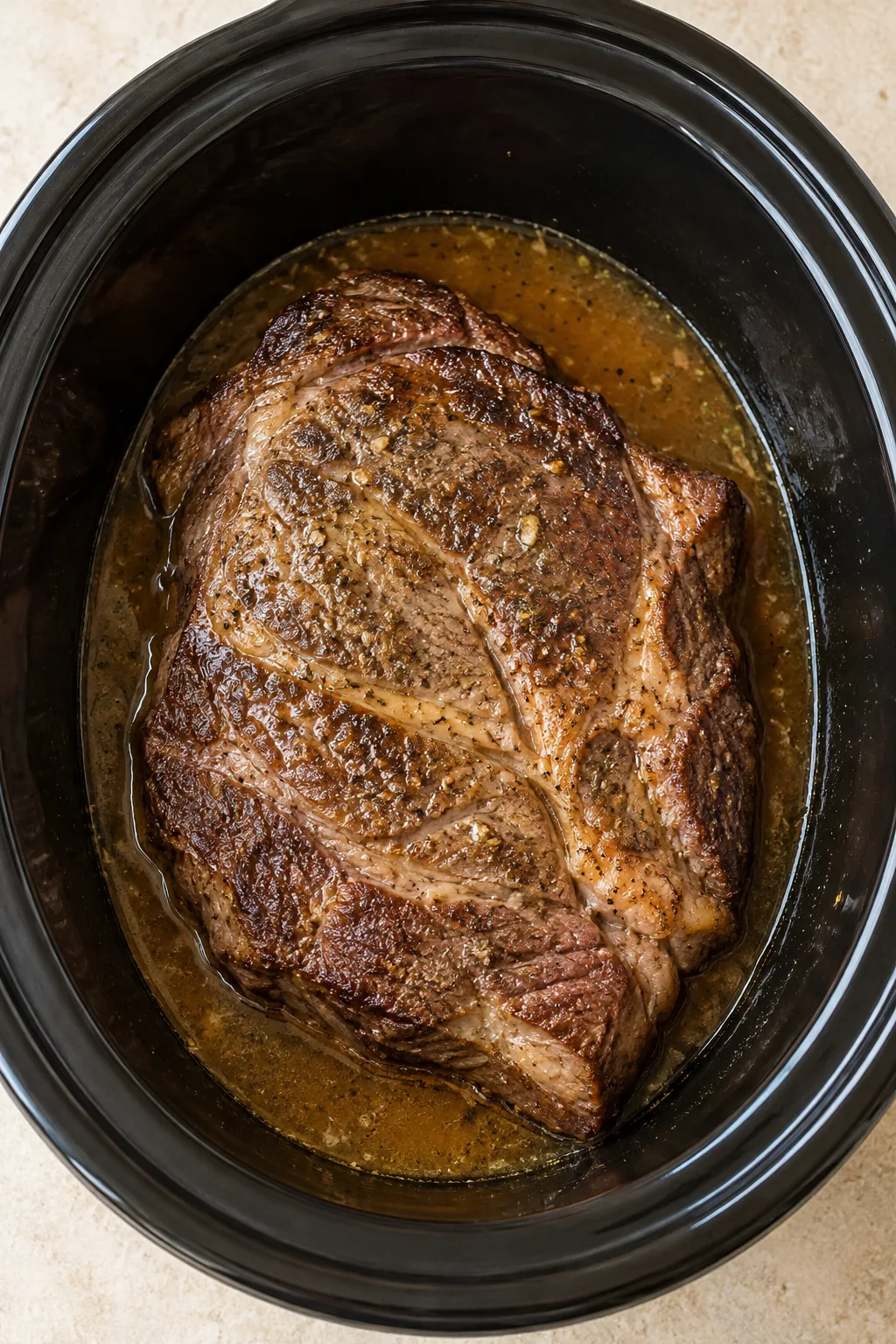 A seared chuck roast with seasoned cooking liquid in a black slow cooker
