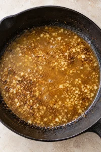 Beef broth and minced garlic loosening browned steak drippings in the skillet.