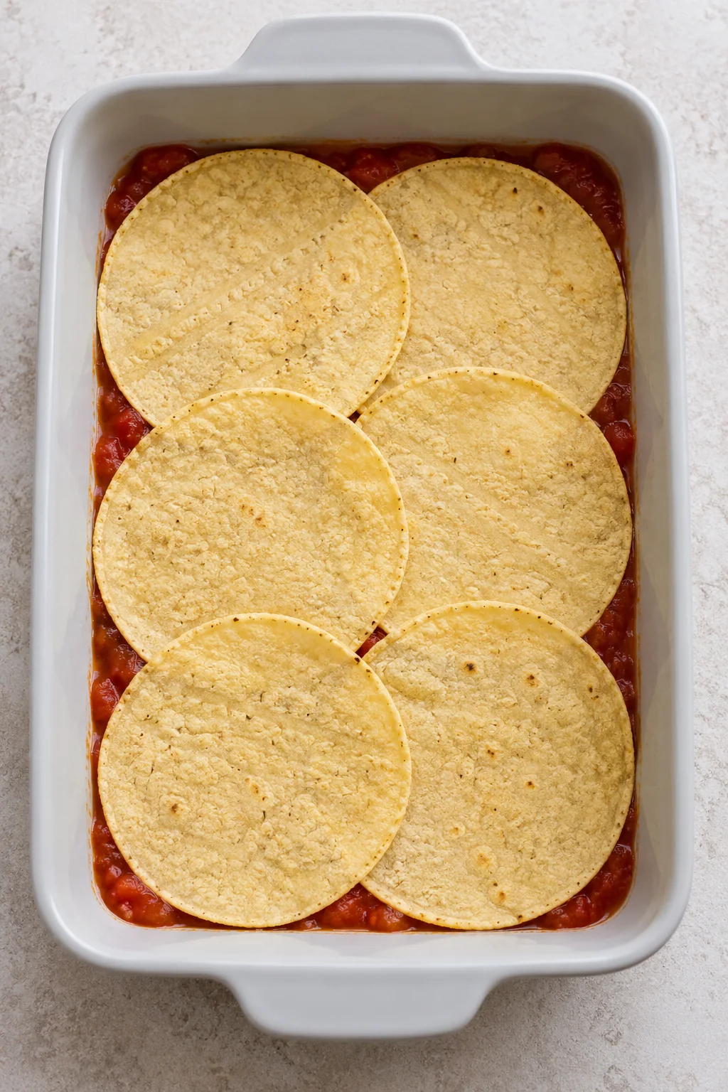 Six corn tortillas overlapping over salsa in a baking dish