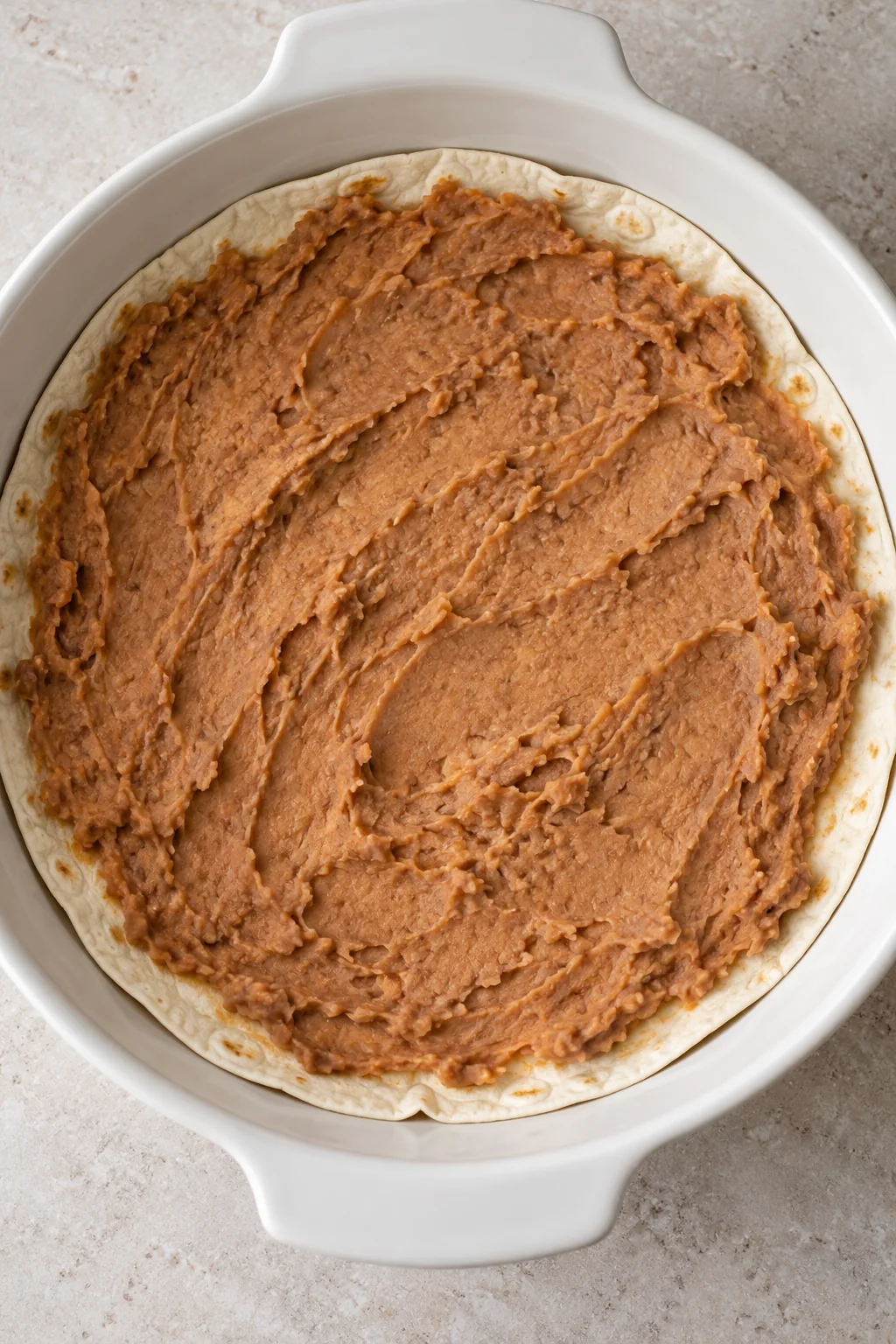 A soft flour tortilla in a round baking dish spread with an even layer of refried beans