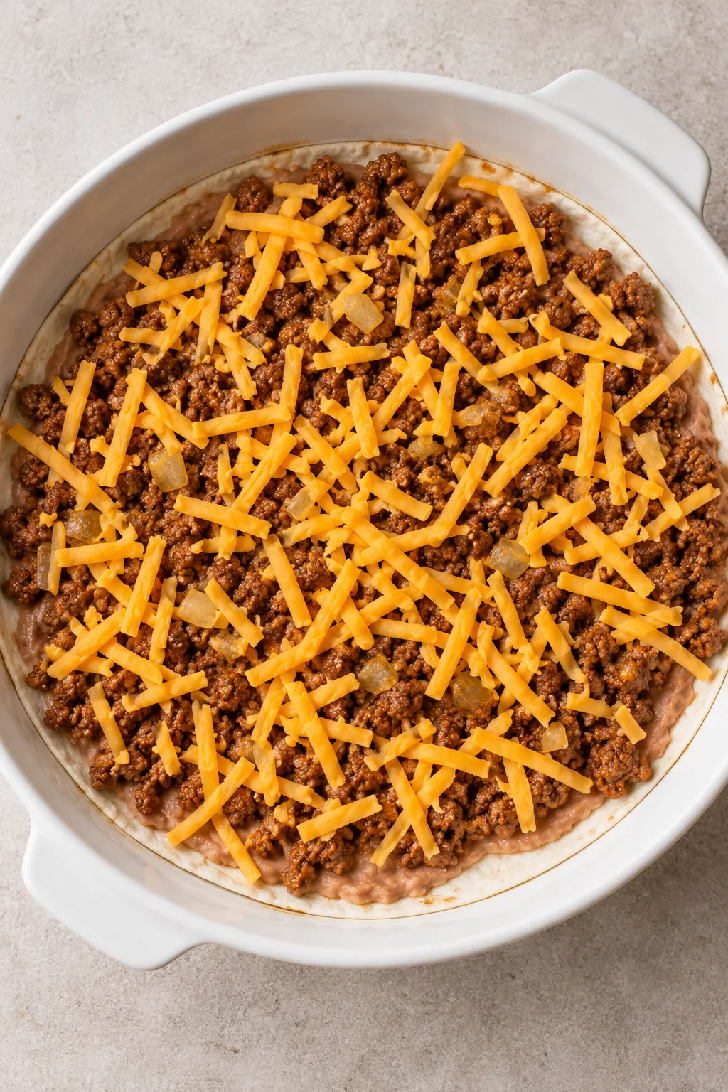 Refried beans, seasoned ground beef and shredded cheddar covering the first tortilla layer