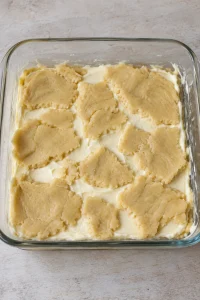 Flattened pieces of raw sugar cookie dough over cream cheese filling in an 8-inch baking dish